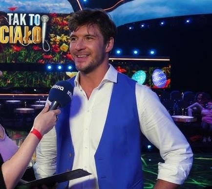 Mikołaj Roznerski na scenie teatralnej w dynamicznej pozie, oświetlony teatralnym światłem, ukazujący jego ekspresję i obecność sceniczną.