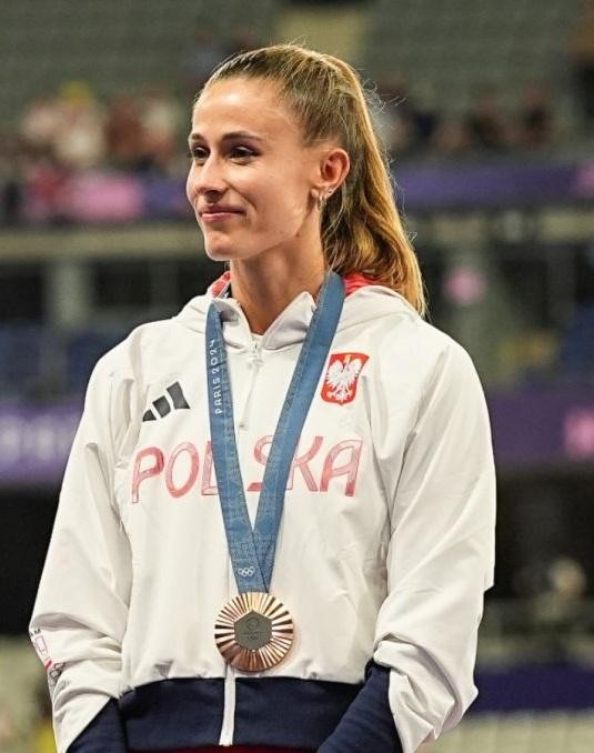 Natalia Bukowiecka uśmiechnięta, przeglądająca Instagram na telefonie, z zamazanym tłem obiektu sportowego.