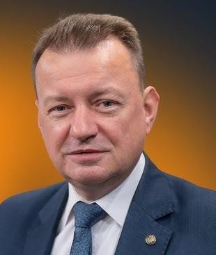 Minister Mariusz Błaszczak podczas ćwiczeń wojskowych, w tle czołgi i żołnierze.
