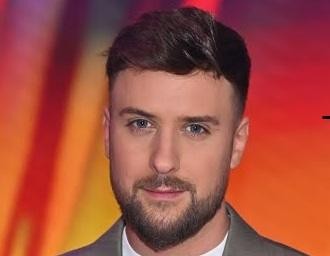 Aleksander Sikora, prezenter telewizyjny, uśmiecha się, przeglądając lub używając telefon komórkowy, co symbolizuje jego aktywność na Instagramie.