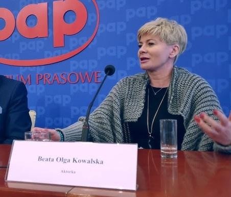 Beata Olga Kowalska na scenie teatralnej, w dynamicznej pozie, oświetlona reflektorem, w kostiumie nawiązującym do jej ról.