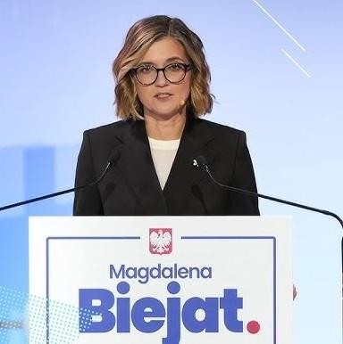 Alt: Magdalena Biejat angażująca się w media społecznościowe, przeglądająca telefon, z tłem sugerującym jej aktywność publiczną.